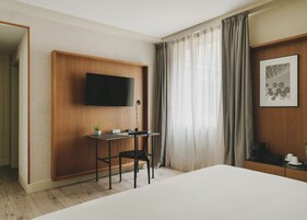 Quarto de hotel com escrivaninha de madeira, televisão de tela plana, uma pequena planta e uma cama.