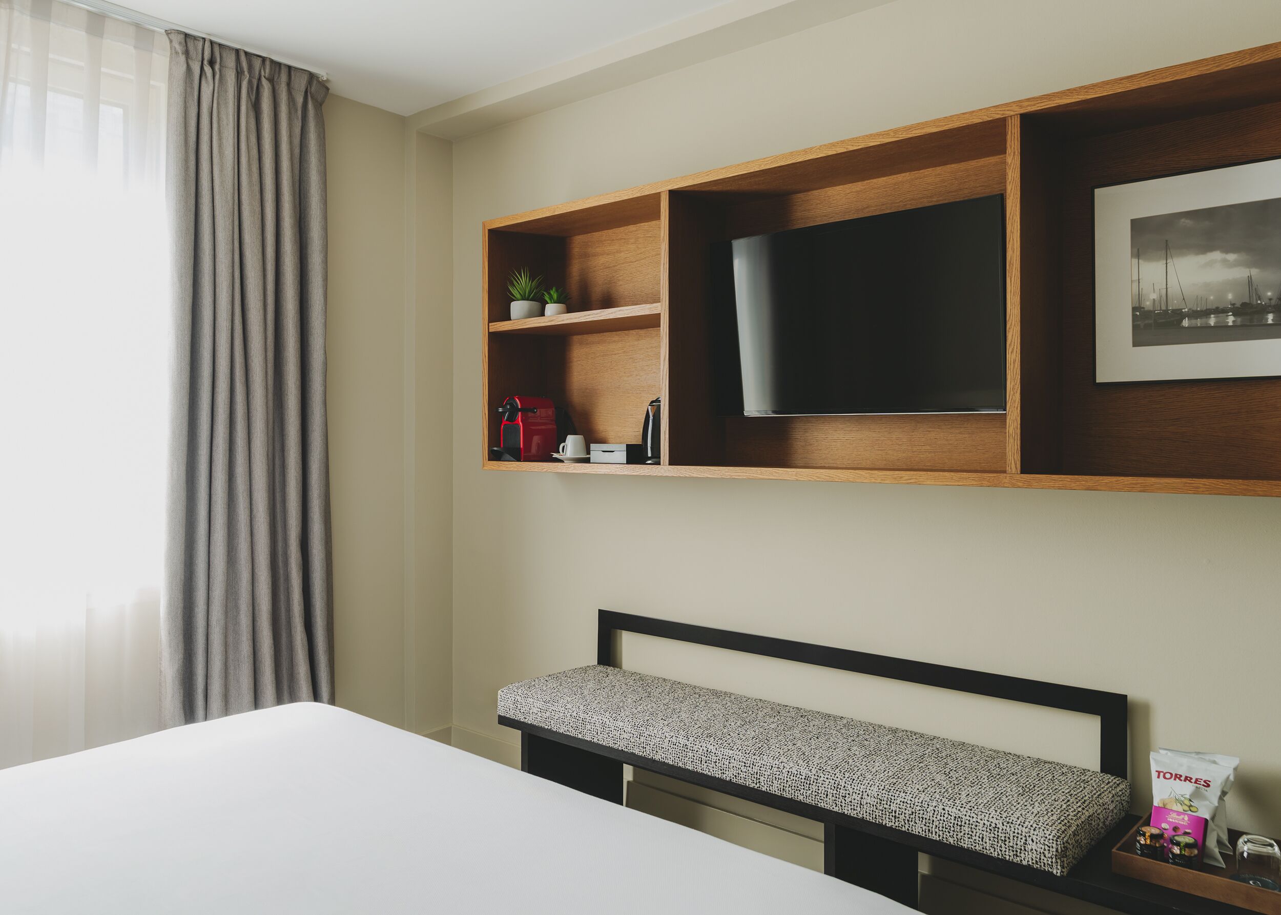 A hotel room with a bed, a TV mounted on a wooden shelf, a small plant, and a framed picture on the wall.