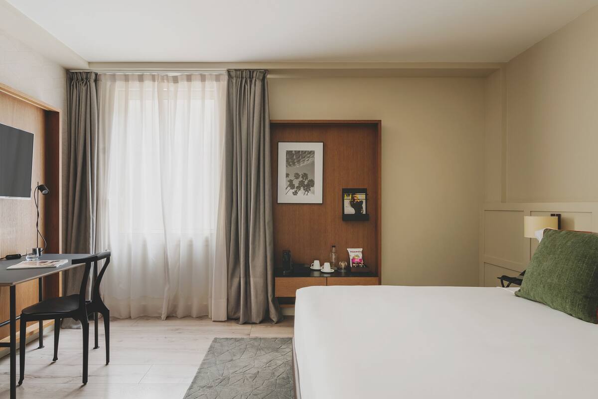 A hotel room with a bed, a desk with a chair, a TV, and a bedside table with a lamp and a picture frame.