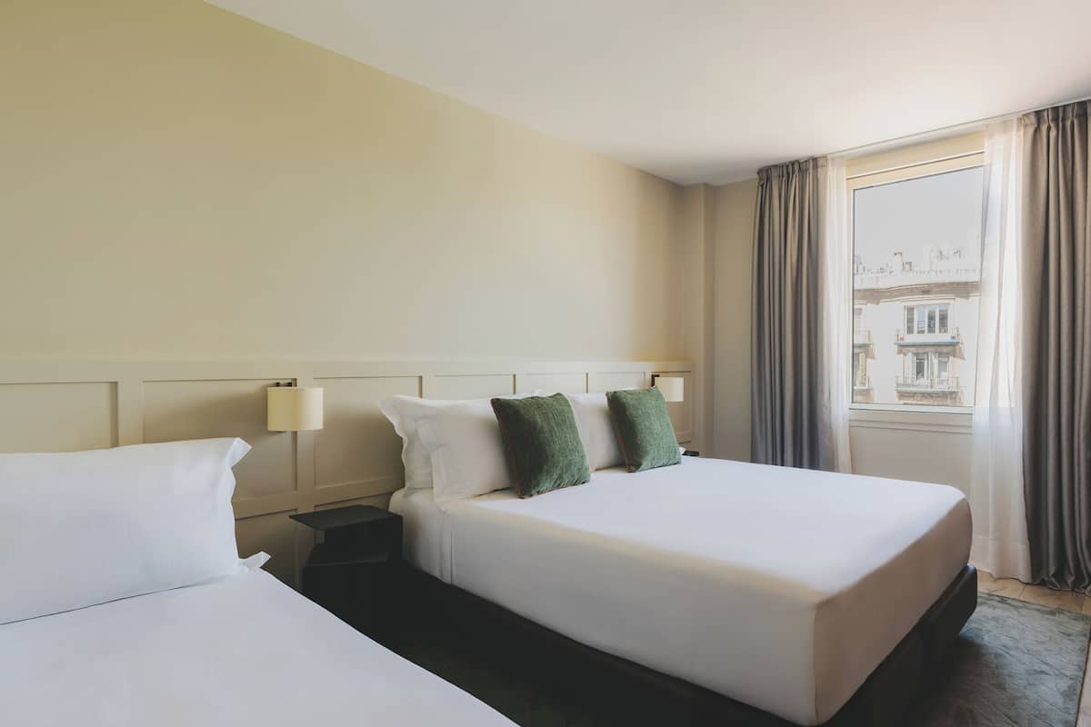 A hotel room with two beds, a nightstand, a lamp, and a window with curtains.