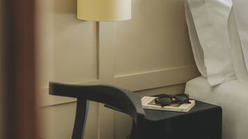 Habitación de hotel impecable, con una mesita de noche que sostiene un libro y gafas, una lámpara y una toalla blanca doblada con cuidado.