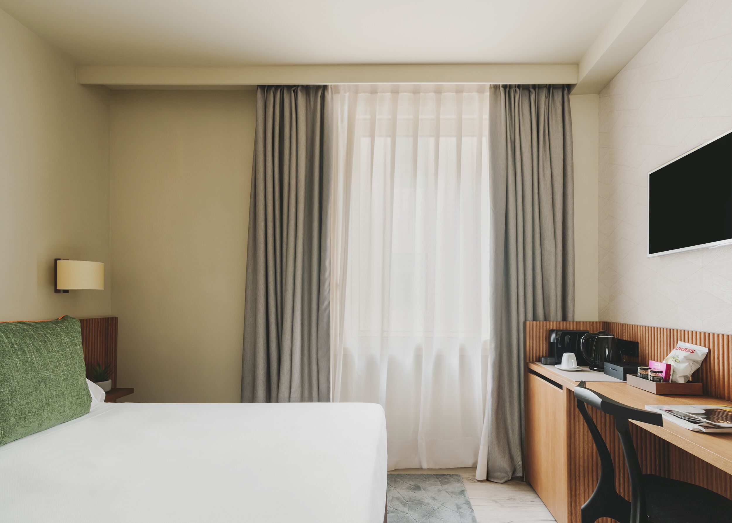 Habitación de hotel con cama, escritorio con silla, cafetera, televisor y ventana con cortinas.