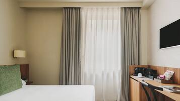 Habitación de hotel con cama, escritorio con silla, cafetera, televisor y ventana con cortinas.