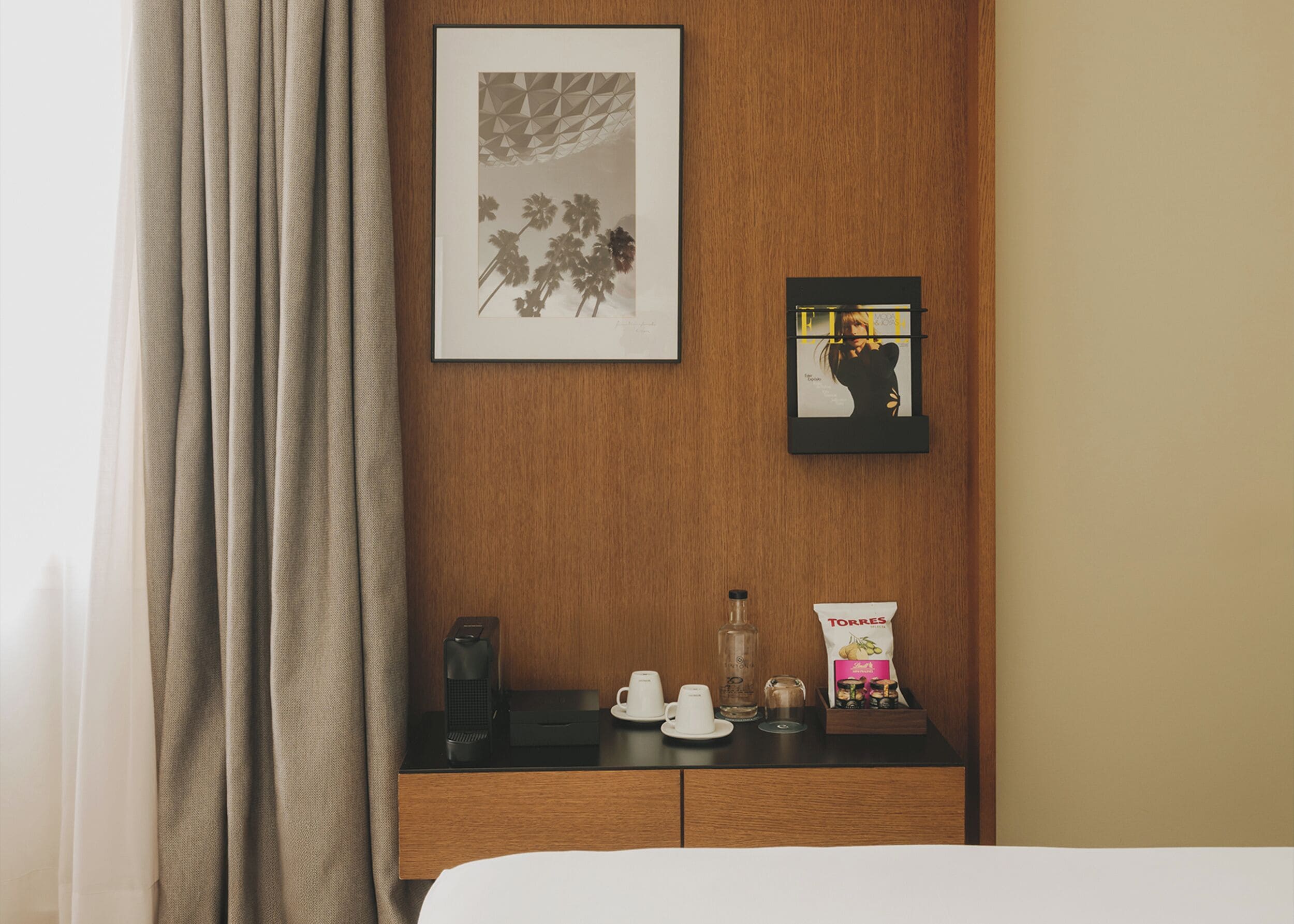 Une chambre d’hôtel avec un mur recouvert de panneaux en bois, un tableau représentant une plante, une photographie encadrée d’une personne, un mini-bar avec une machine à café, une bouteille et un paquet de collations.