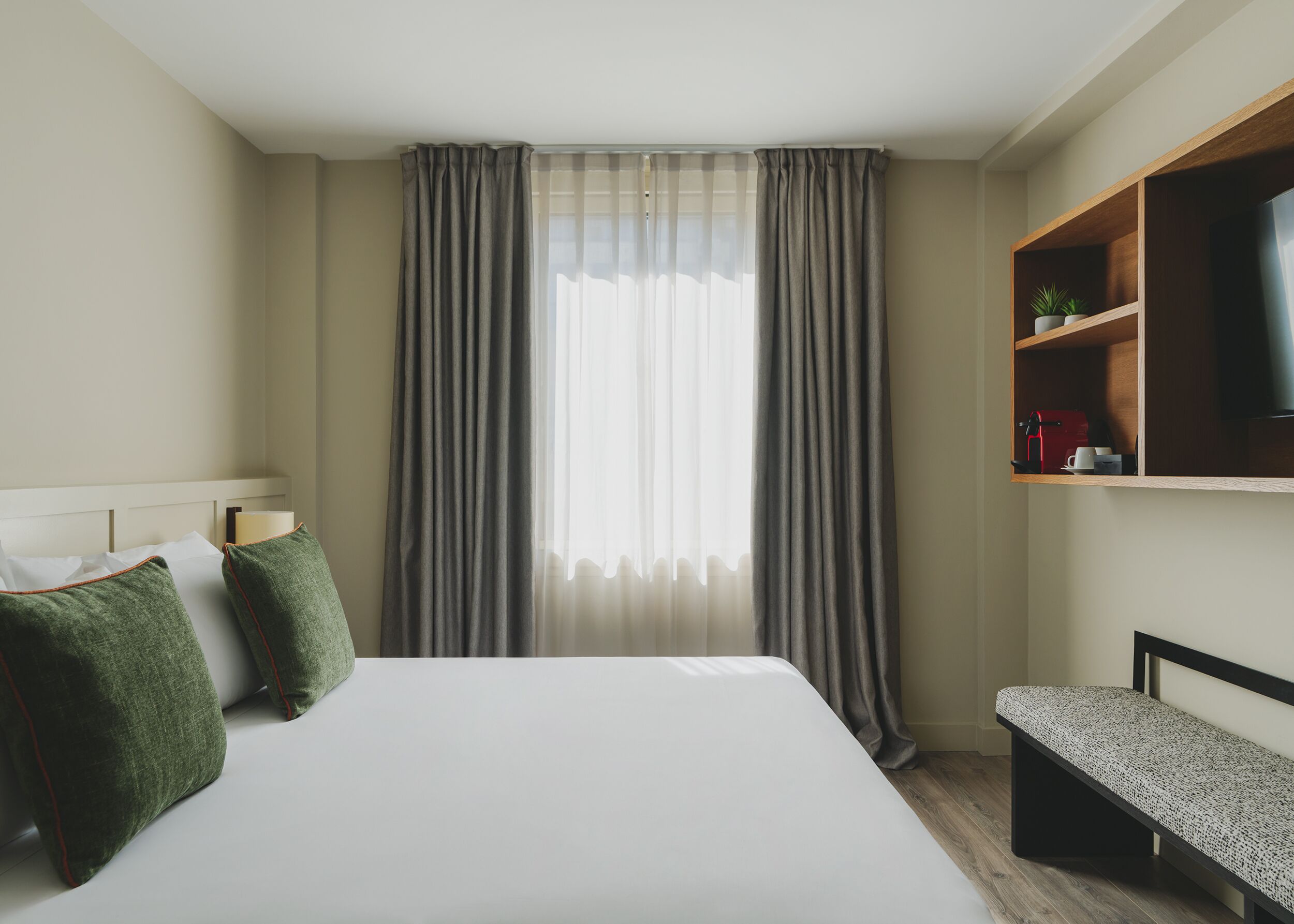 Quarto de hotel com uma cama grande, cortinas cinzas, uma estante com uma TV e um banco.