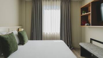 Una habitación de hotel con una cama grande, cortinas grises, un estante de madera con un televisor y un banco.