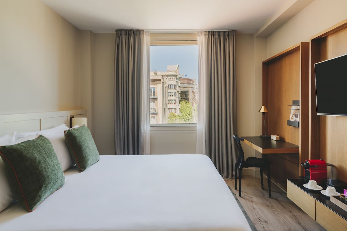 A hotel room with a bed, a desk, a chair, a TV, and a window with curtains.