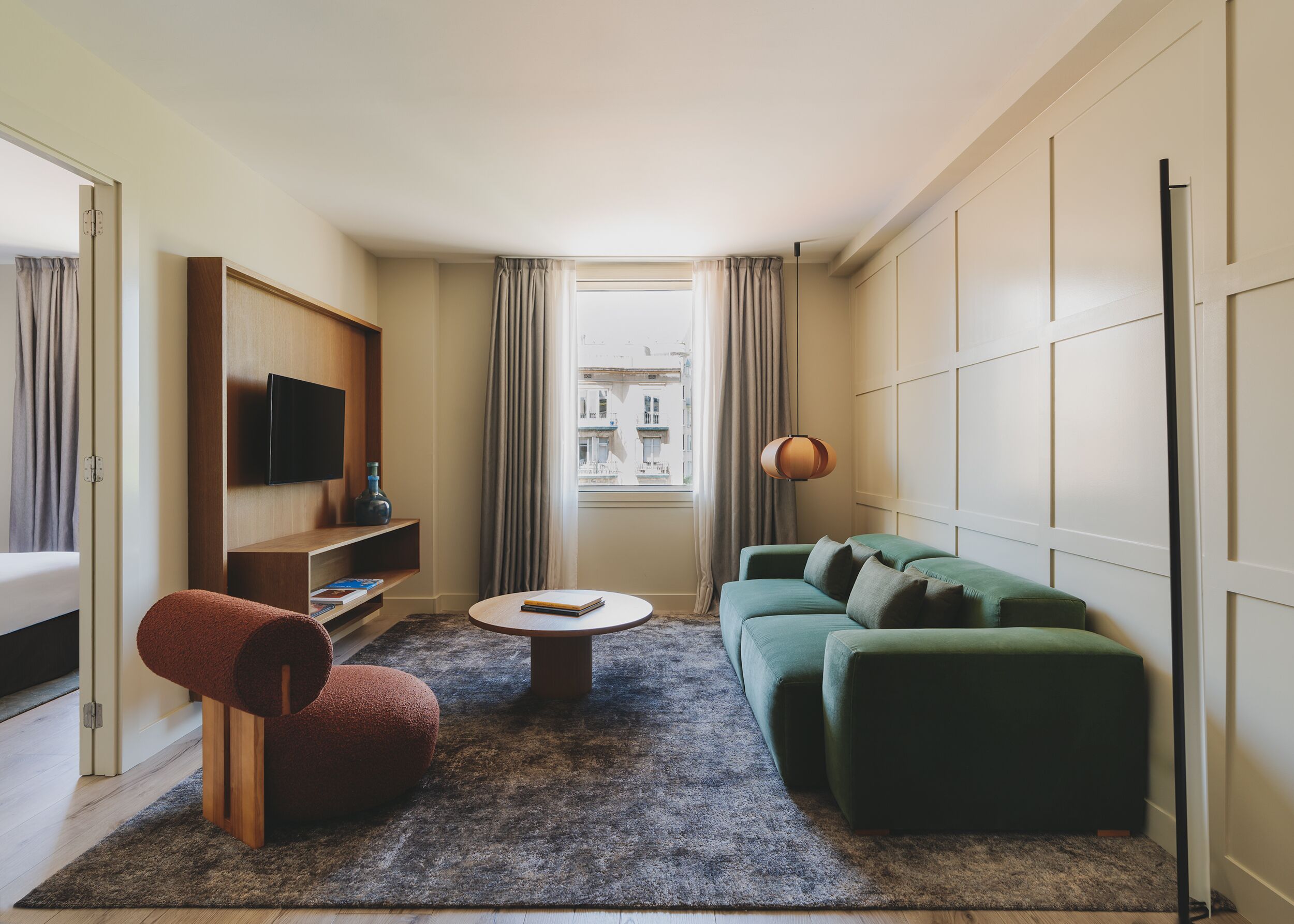 Habitación de hotel compacta con sofá verde, una mesita redonda pequeña, una silla roja, un televisor en un estante de madera y vistas a edificios por la ventana.