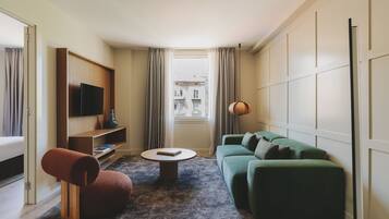 Habitación de hotel compacta con sofá verde, una mesita redonda pequeña, una silla roja, un televisor en un estante de madera y vistas a edificios por la ventana.