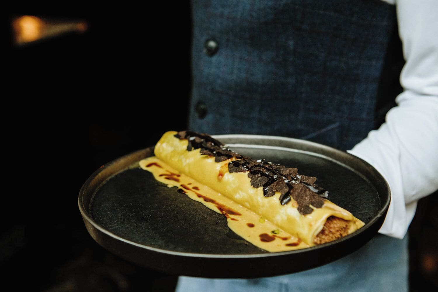 A person holding a tray with a rolled dessert, possibly a crepe or pastry, topped with chocolate shavings and sauce.