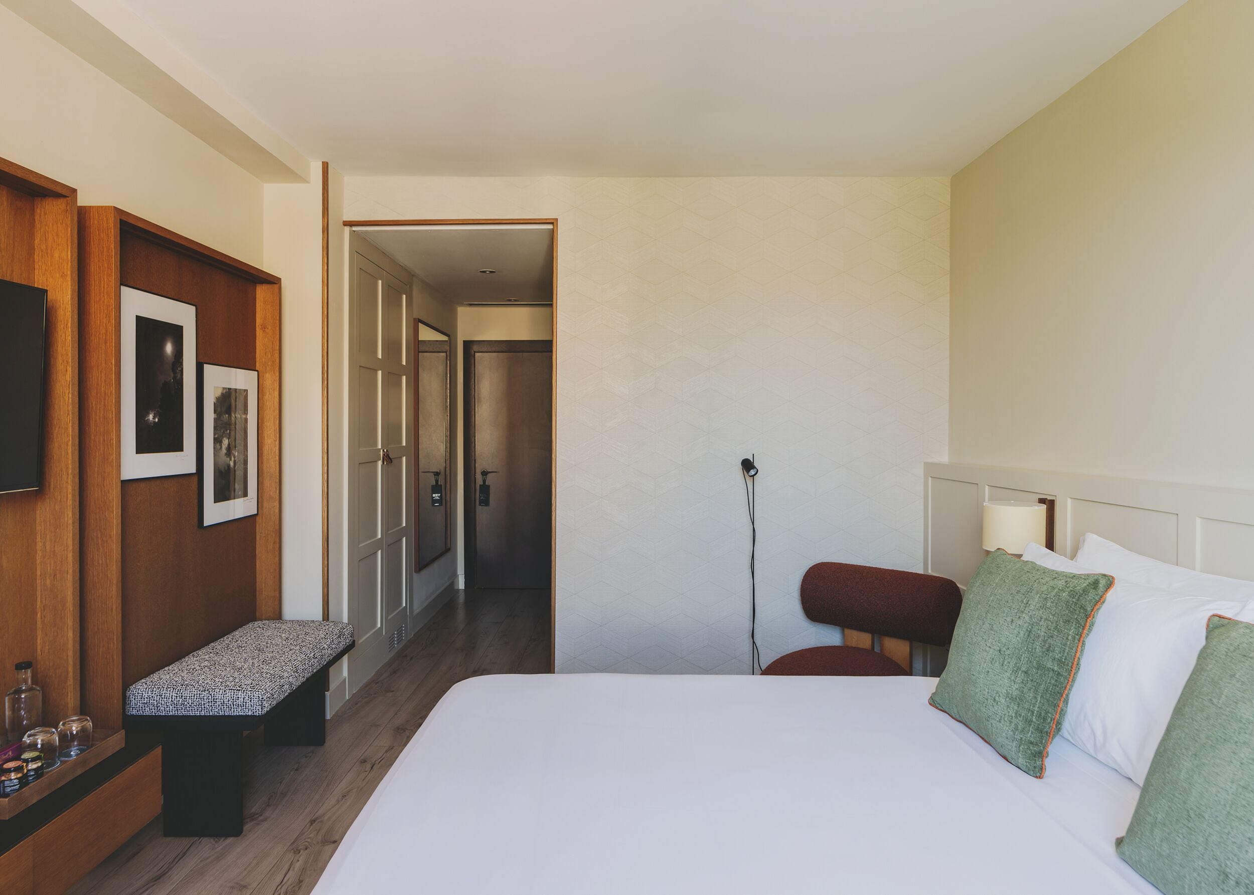 Habitación de hotel con una cama grande, un banco, un escritorio con lámpara y un televisor de pared.