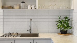 Una cocina con un lavamanos de acero inoxidable, salpicadero de azulejos blancos y una planta de bambú en maceta.