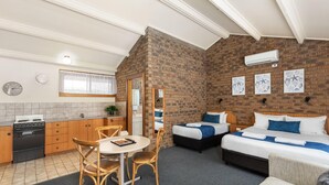 Habitación de hotel compacta con área de cocina, dos camas, una mesa de comedor con sillas y una pared decorativa de ladrillo.