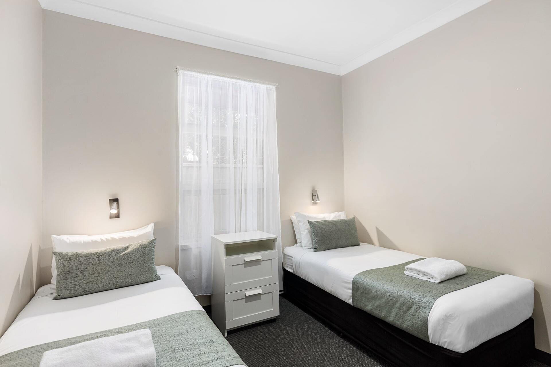 Habitación de hotel con dos camas individuales, un tocador blanco, una ventana con cortinas traslúcidas e iluminación empotrada.