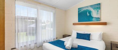 A hotel room with a bed, a painting of a wave, and a window with sheer curtains.