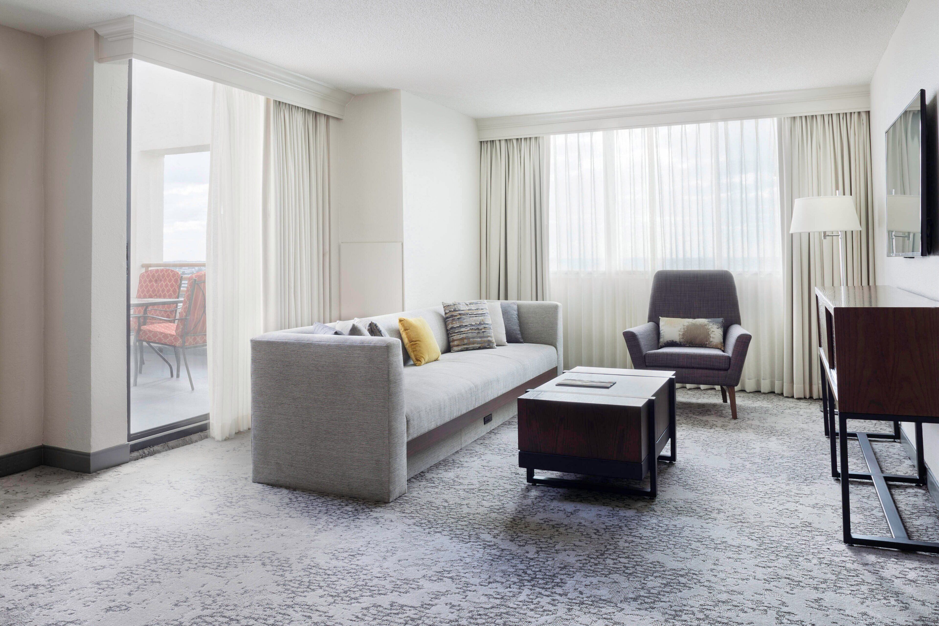 Habitación de hotel moderna con un sofá gris, un sillón pequeño, una mesa de centro de madera y un gran ventanal con cortinas.