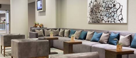 A modern lobby with a grey sectional sofa, multiple armchairs, and small wooden tables with plants.