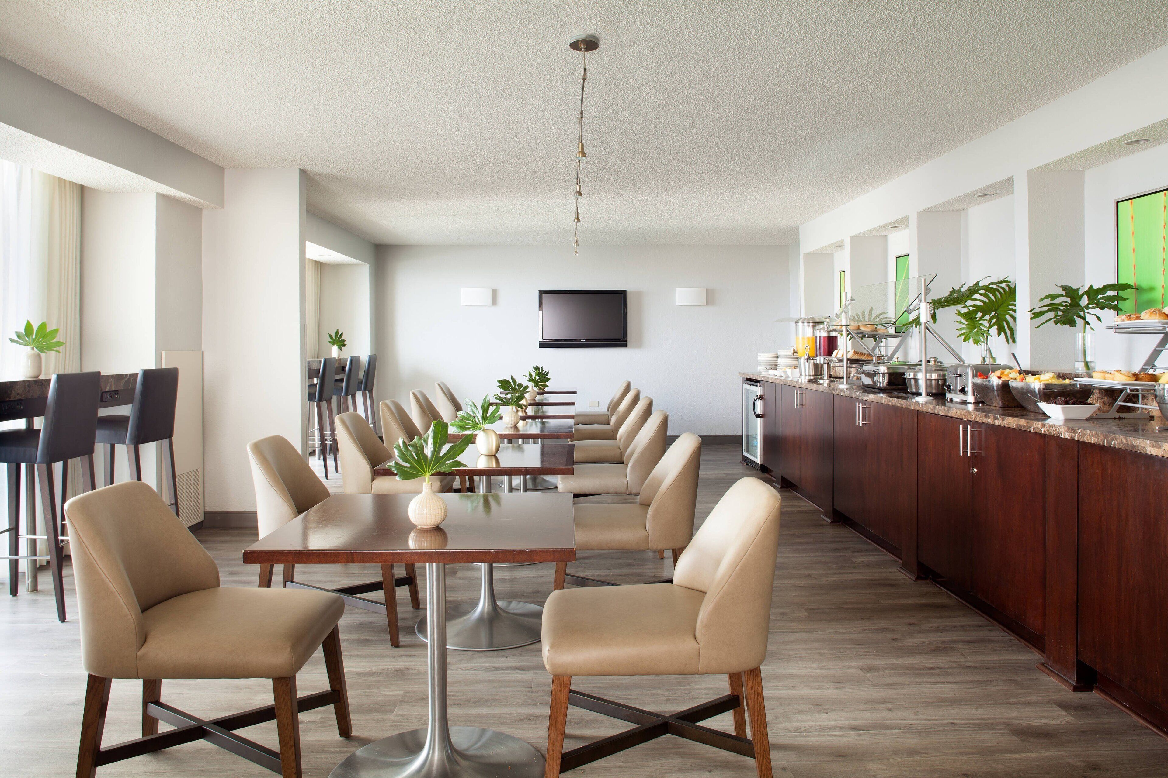 A modern kitchen with a dining area, featuring a long table with chairs, a kitchen counter with appliances, and a TV mounted on the wall.