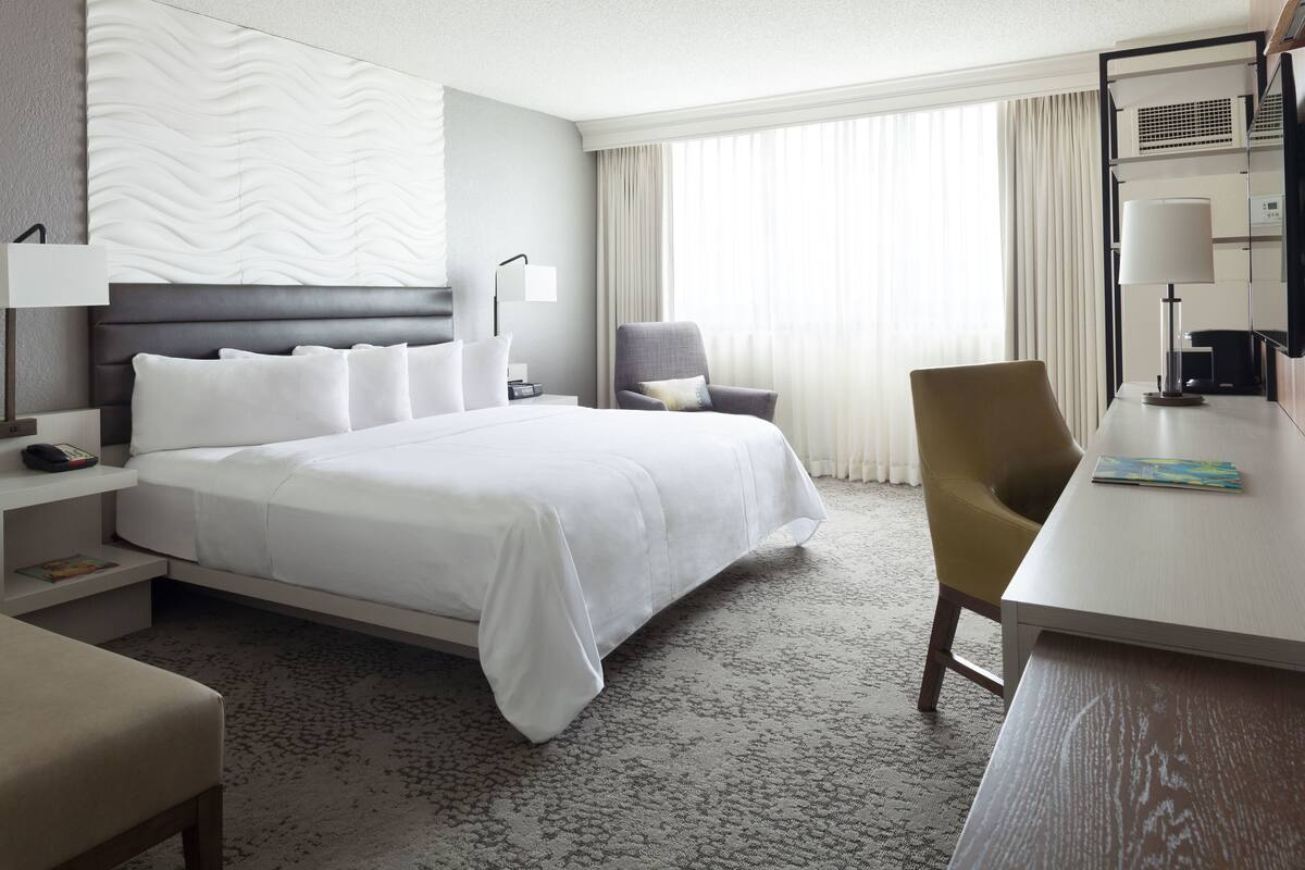 Ein Hotelzimmer mit einem großen Bett, einem Schreibtisch, einem Stuhl, einer Lampe und einem Vorhangfenster.