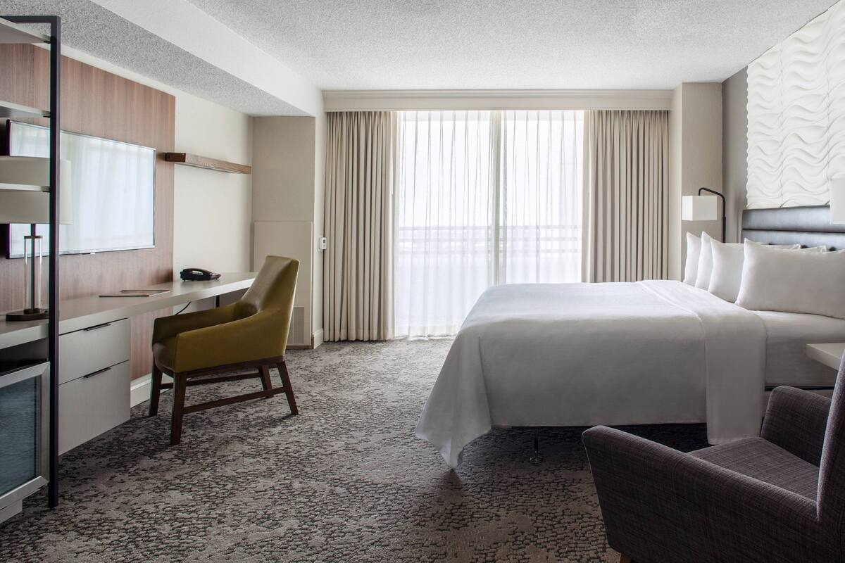 Ein Hotelzimmer mit Bett, Schreibtisch, Stuhl und einem großen Fenster mit Vorhängen.