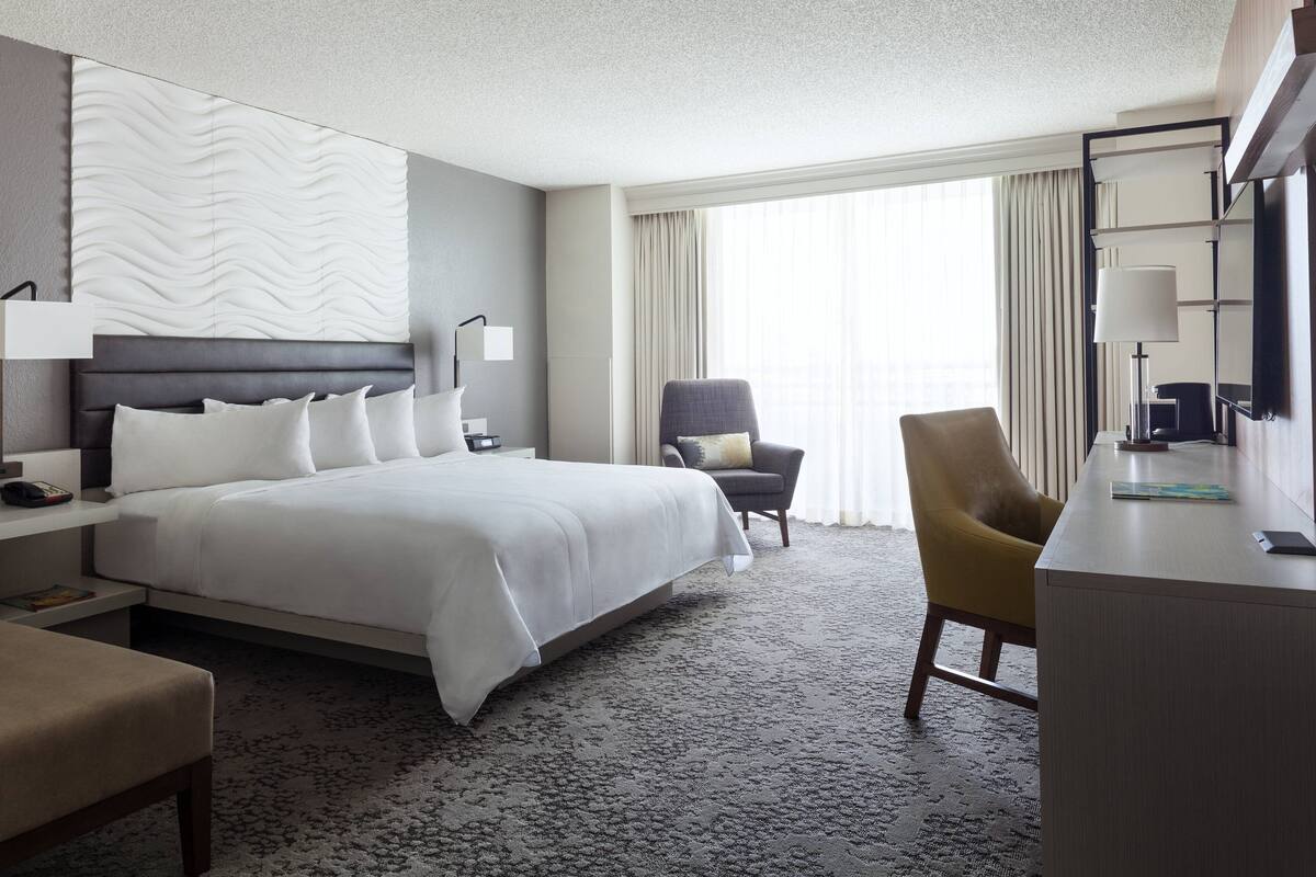 Ein Hotelzimmer mit einem großen Bett, einem Schreibtisch, einem Sessel und Blick ins Freie durch das Fenster.