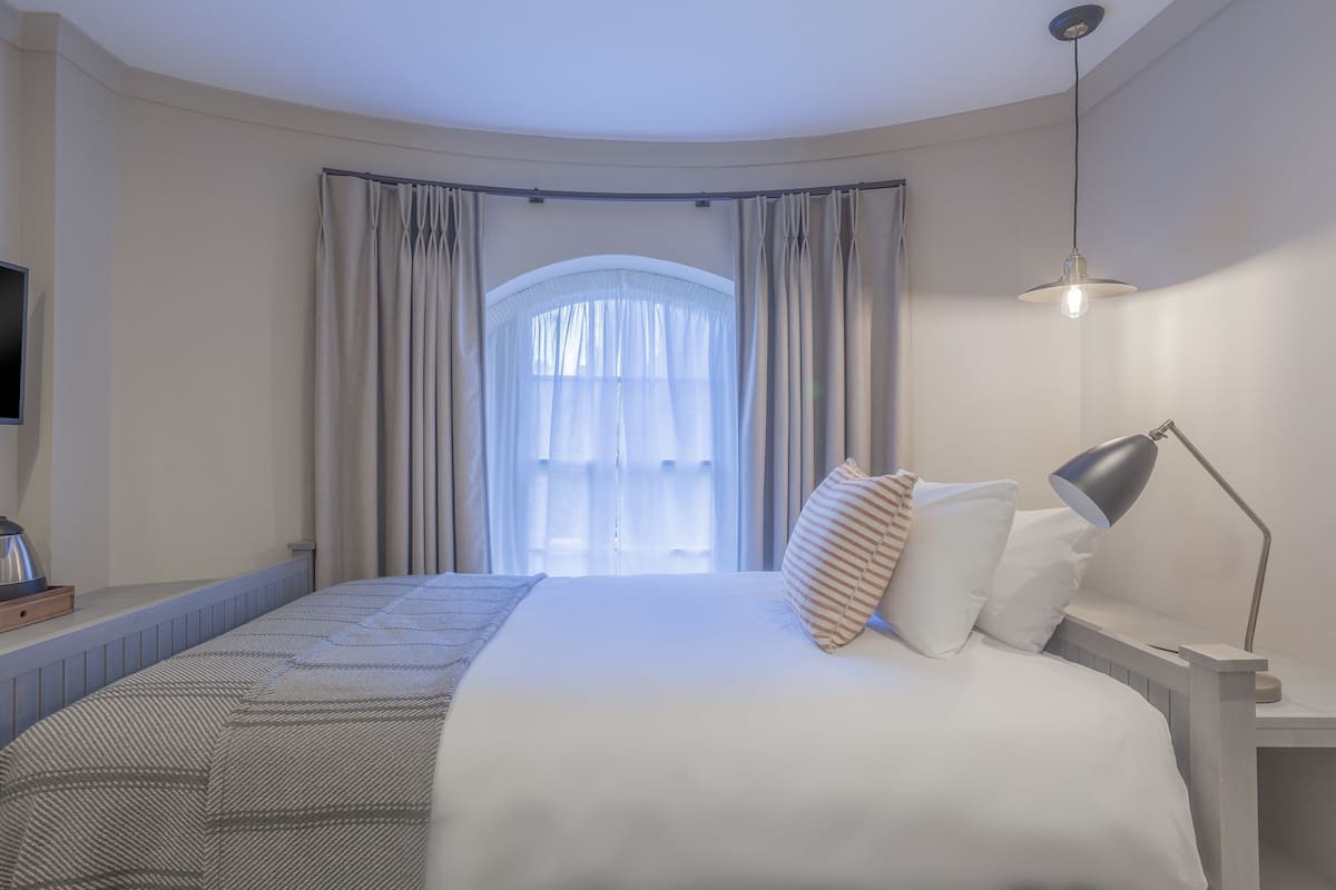 A hotel room with a large bed, a bedside table with a lamp, a TV on the left, and a window with curtains.