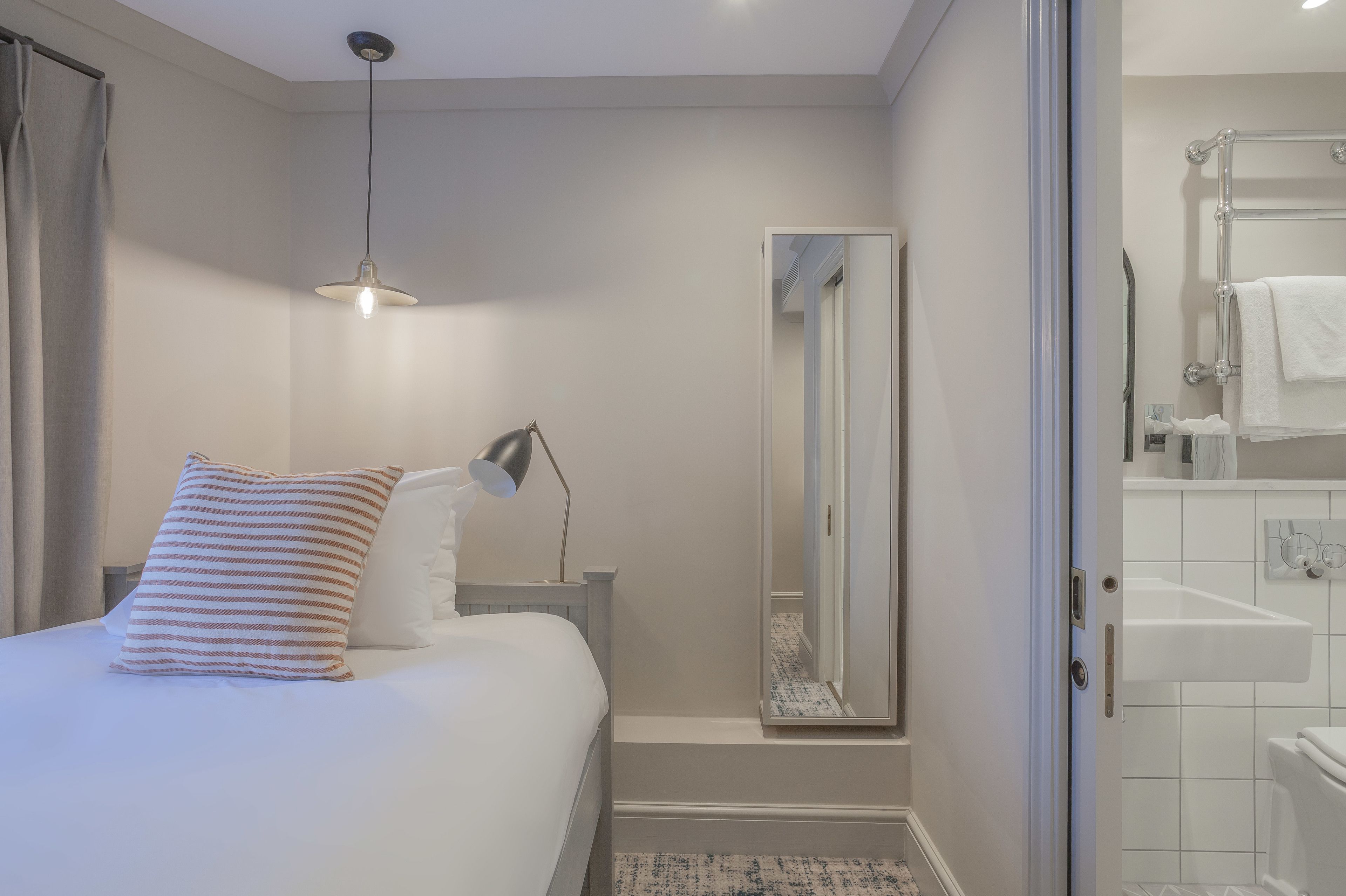 A bedroom with a bed, a bedside lamp, a mirror, and a bathroom visible through an open door.