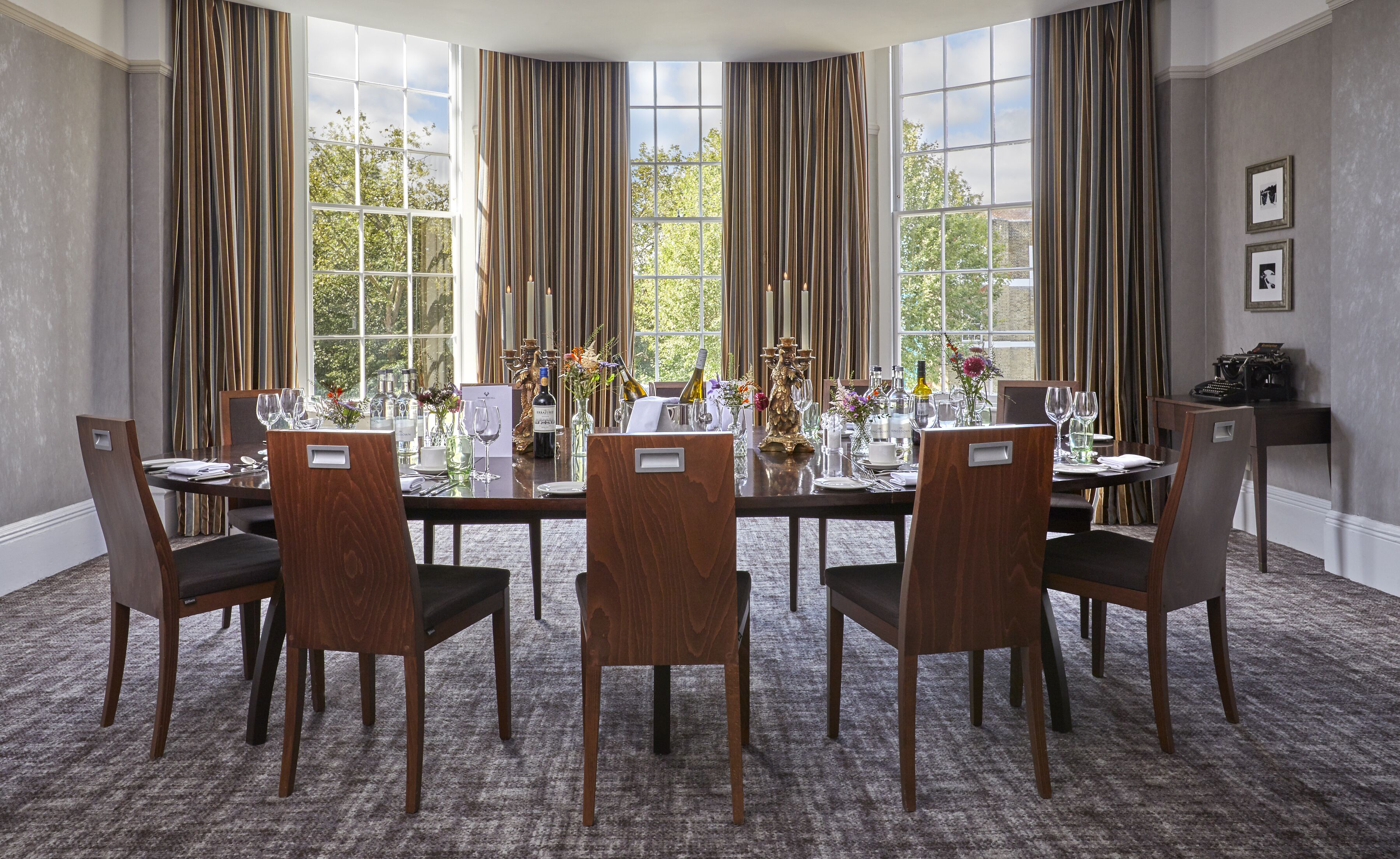 Ein Esszimmer mit einem großen Tisch, gedeckt für sechs Personen, umgeben von hohen Fenstern mit Vorhängen und einem Teppichboden.