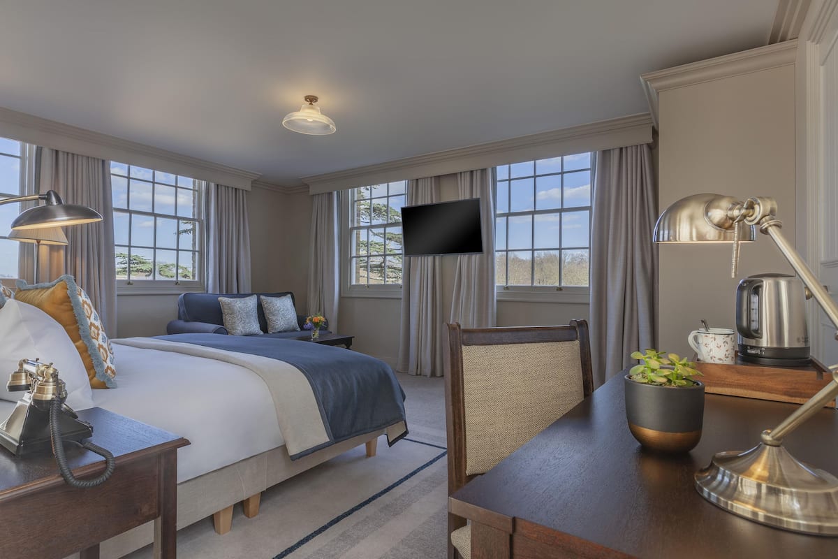 A hotel room with a large bed, a desk with a lamp, a television, and a view of the outdoors through large windows.