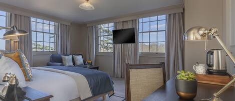A hotel room with a large bed, a desk with a lamp, a television, and a view of the outdoors through large windows.