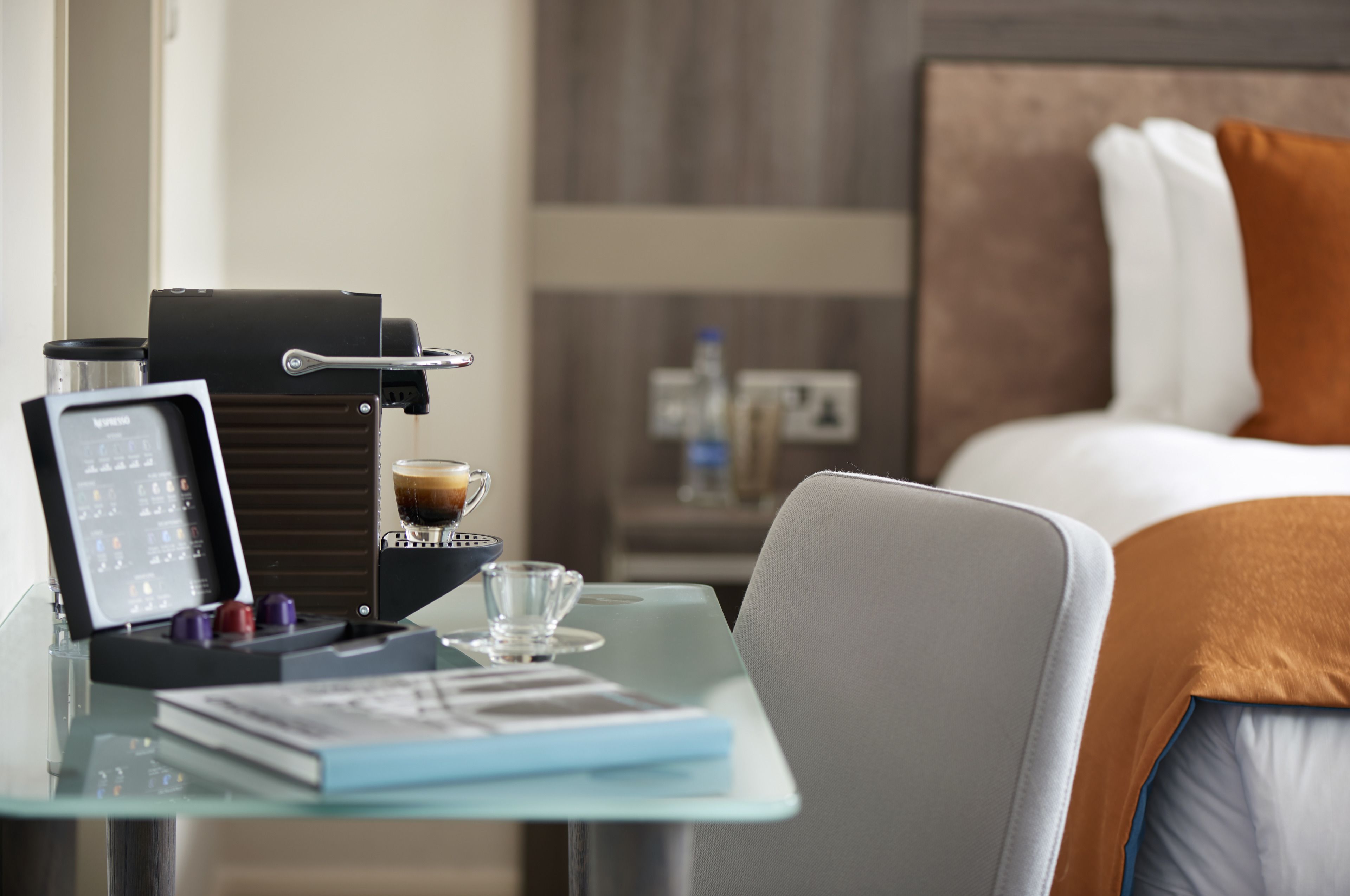 A hotel room with a bed, a glass table with a coffee machine, a laptop, and a book.