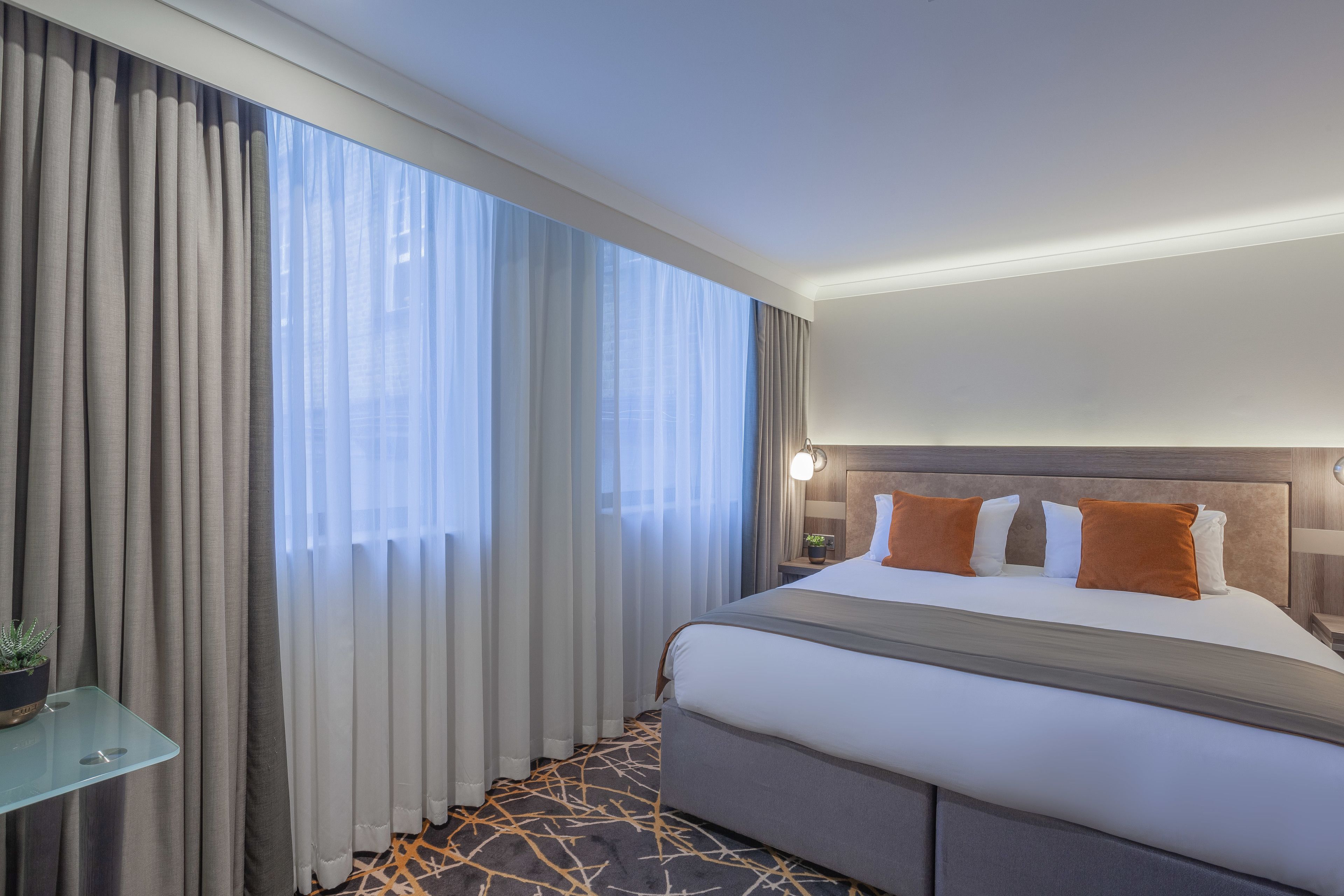 A hotel room with a large bed, a bedside table with a lamp, and a view through sheer curtains.