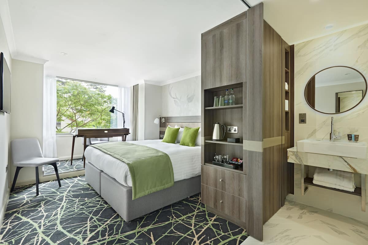 A modern hotel room with a bed, bedside table, chair, wardrobe, and a bathroom with a sink and mirror.