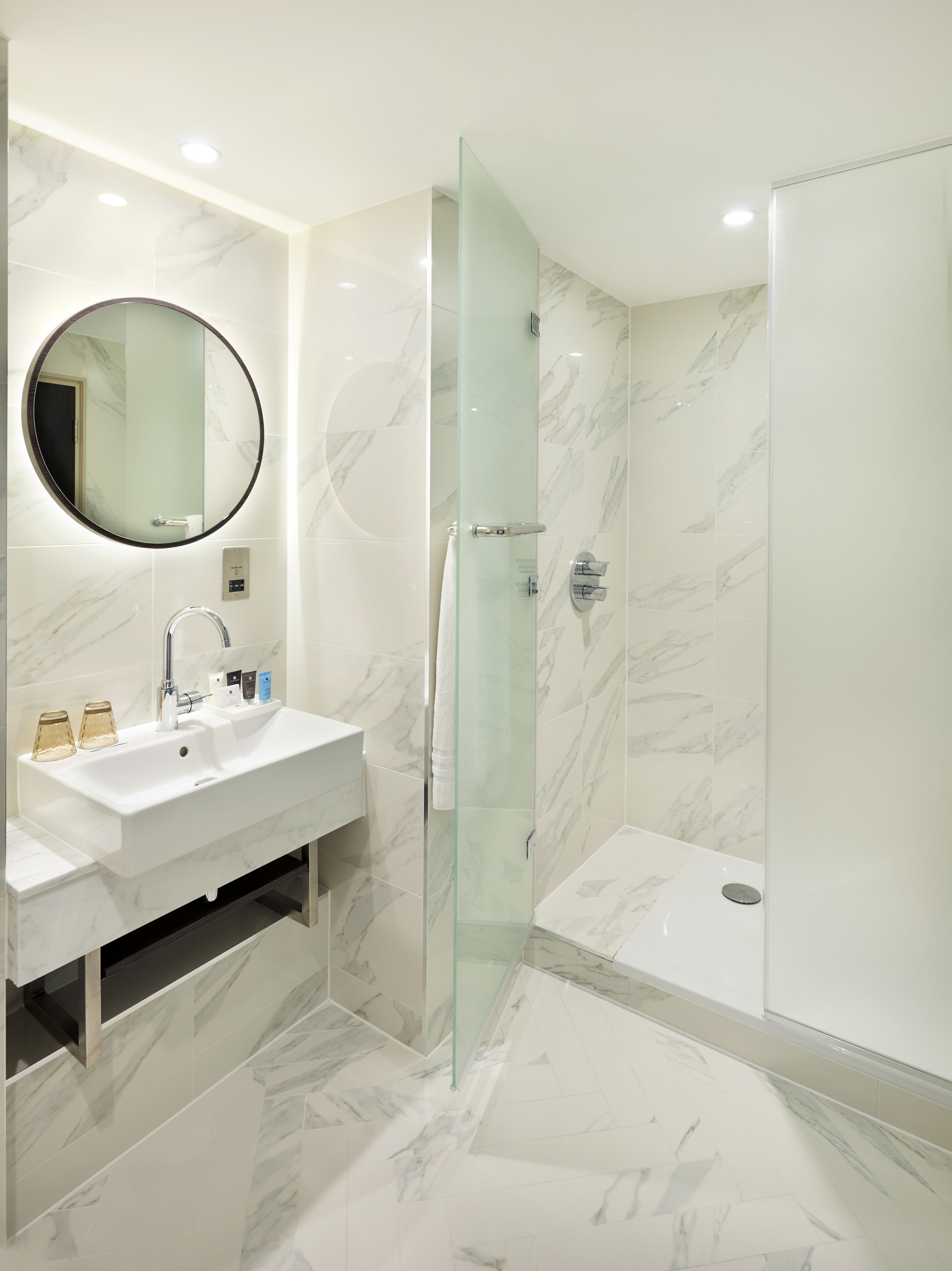 A modern bathroom with a marble sink, a glass shower enclosure, and a round mirror.