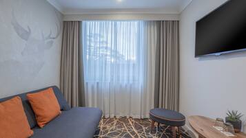 Habitación de hotel moderna con sofá, una pequeña mesa con una planta, televisor y ventana con cortinas.