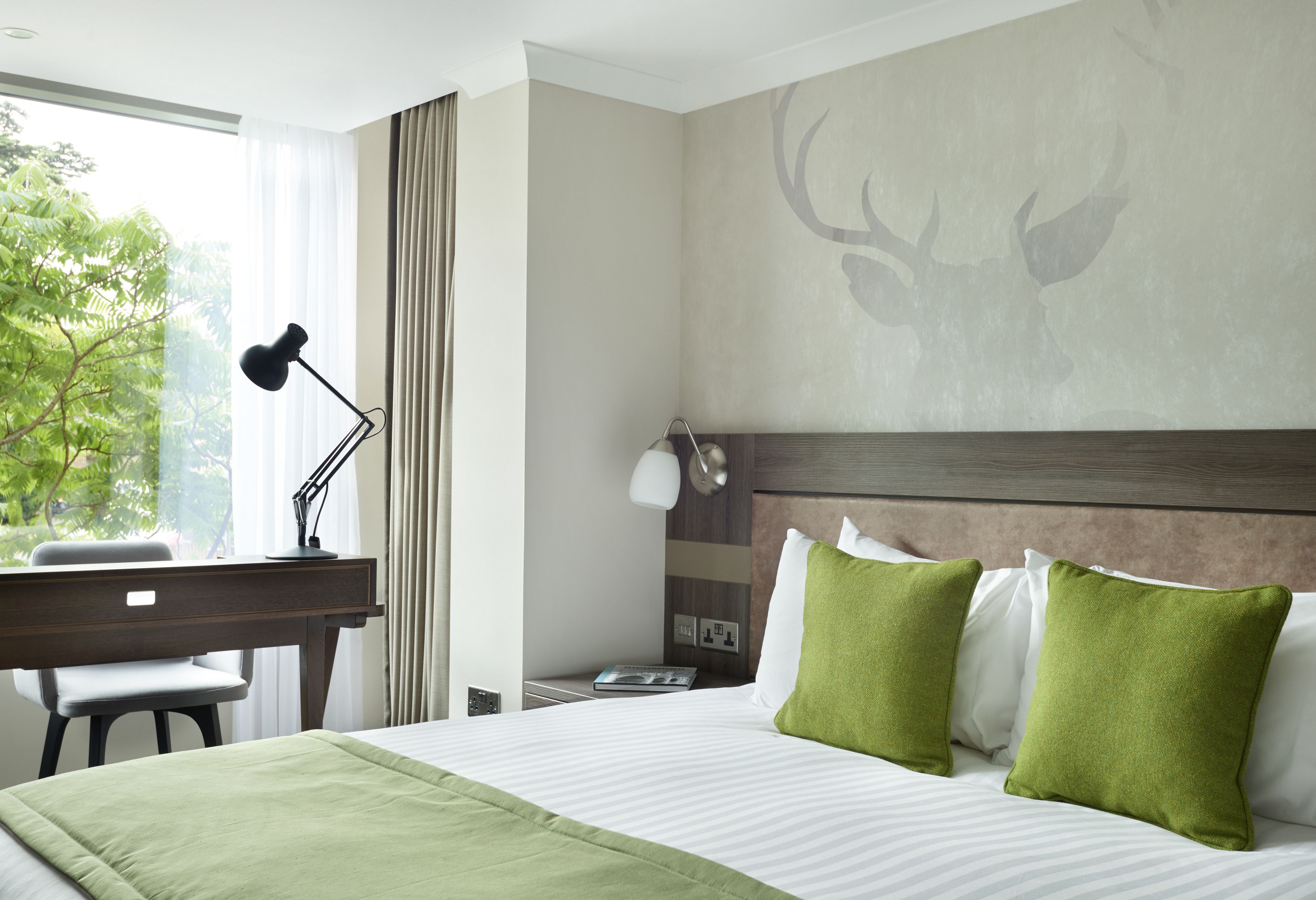 Habitación de hotel con cama, escritorio, silla, lámpara y vistas a zonas verdes.
