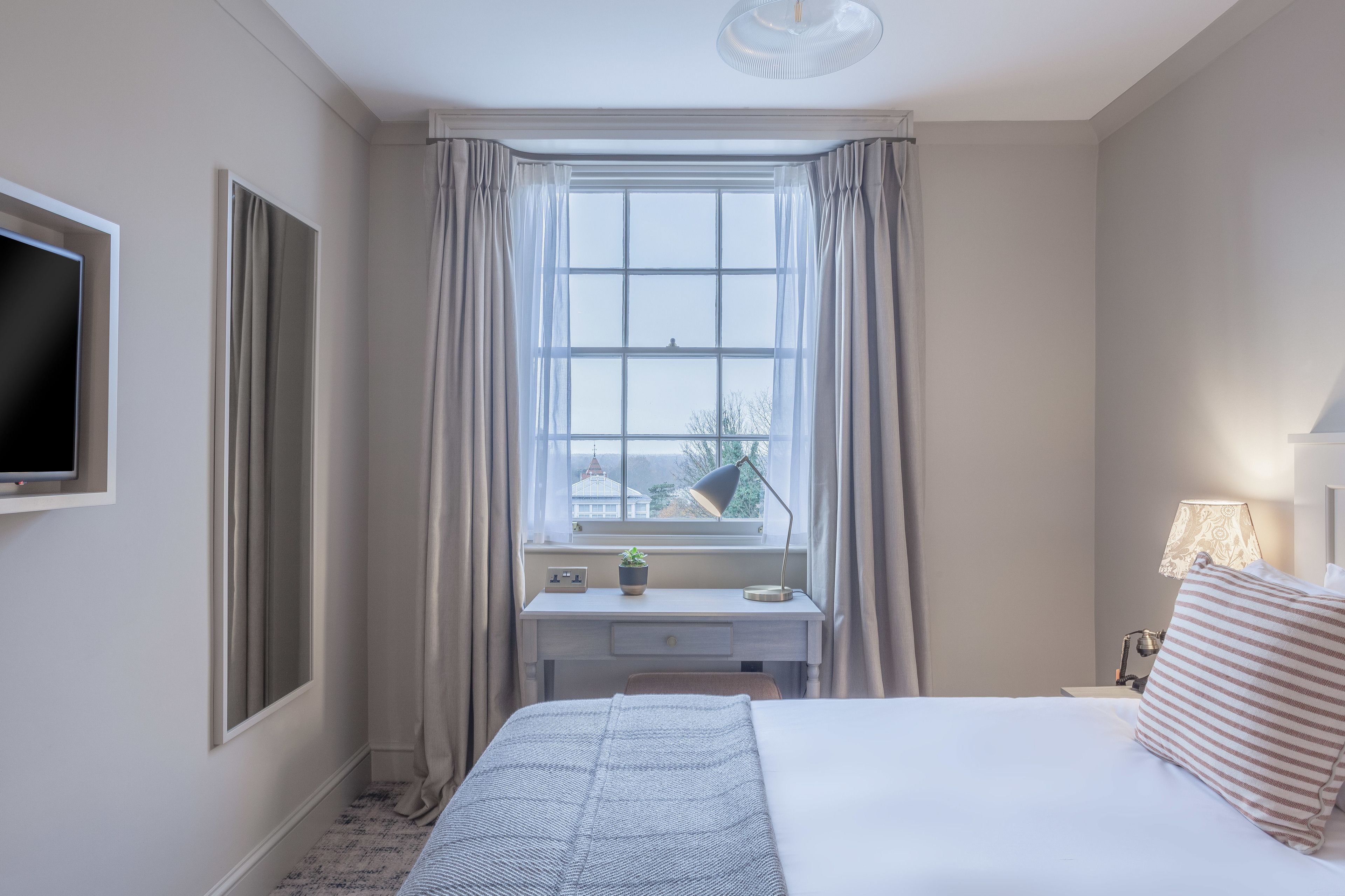 Habitación de hotel con cama, escritorio con lámpara, televisión y ventana con cortinas.