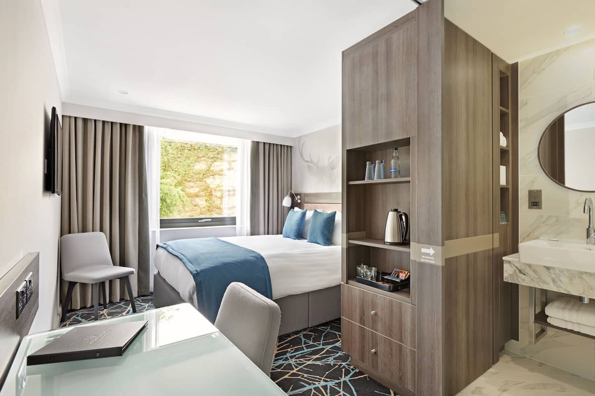 A hotel room with a bed, a desk with a laptop, a chair, a wardrobe, a bathroom with a sink and mirror, and a window with curtains.