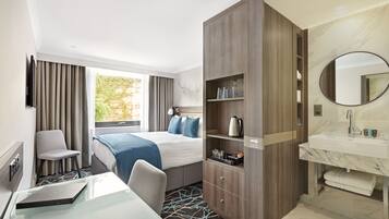 A hotel room with a bed, a desk with a laptop, a chair, a wardrobe, a bathroom with a sink and mirror, and a window with curtains.