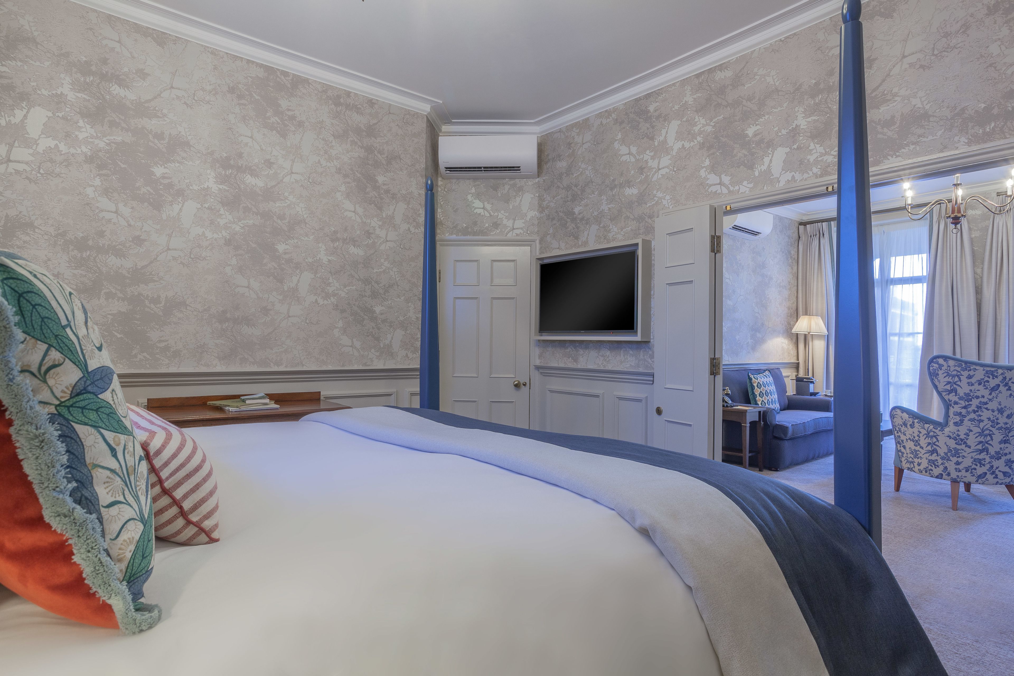 A hotel room with a large bed, a flat-screen TV mounted on the wall, a seating area with a sofa and armchairs, and a door leading to another room.