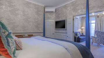 Habitación de hotel con una cama grande, un televisor de pantalla plana montado en la pared, una zona de estar con un sofá y sillones, y una puerta que da a otra habitación.