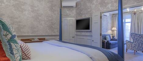 A hotel room with a large bed, a flat-screen TV mounted on the wall, a seating area with a sofa and armchairs, and a door leading to another room.