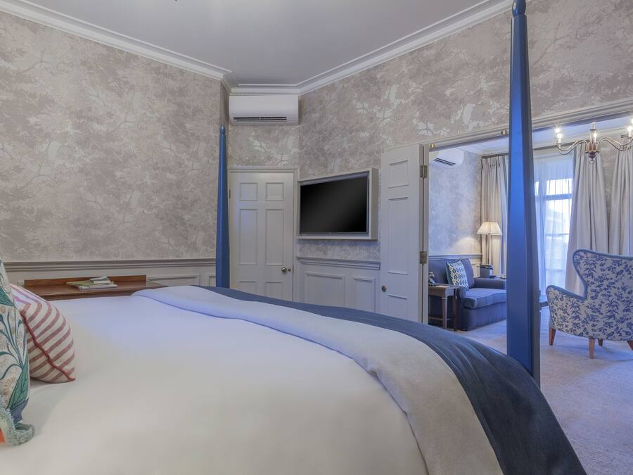 Habitación de hotel con una cama grande, un televisor de pantalla plana montado en la pared, una zona de estar con un sofá y sillones, y una puerta que da a otra habitación.