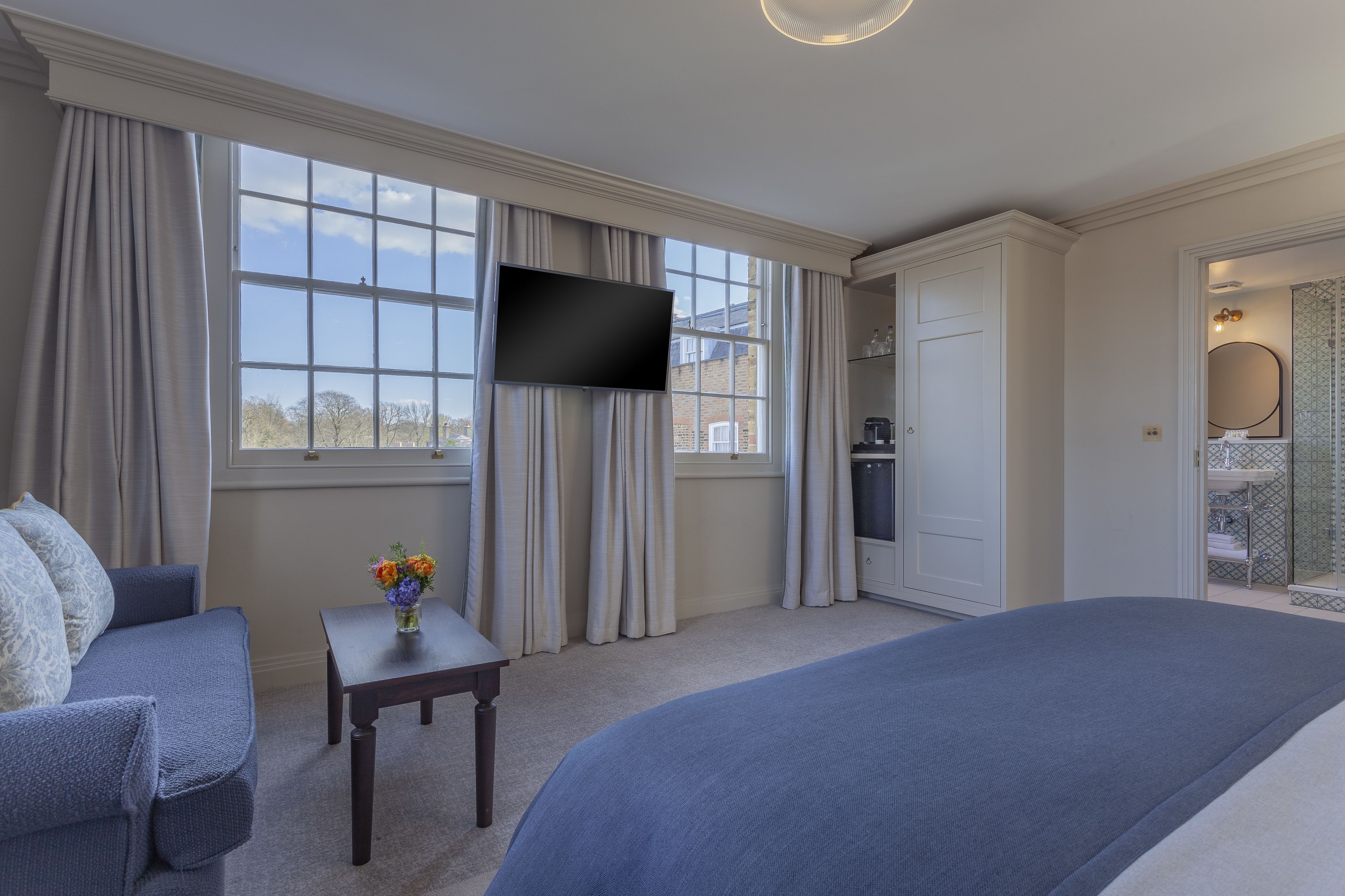 A hotel room with a large bed, a TV mounted on the wall, a seating area with a small table and chairs, and a view of the outside through large windows.