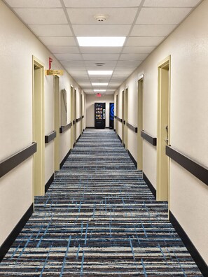 Un pasillo de hotel con piso alfombrado, puertas a ambos lados y una máquina expendedora en el centro.