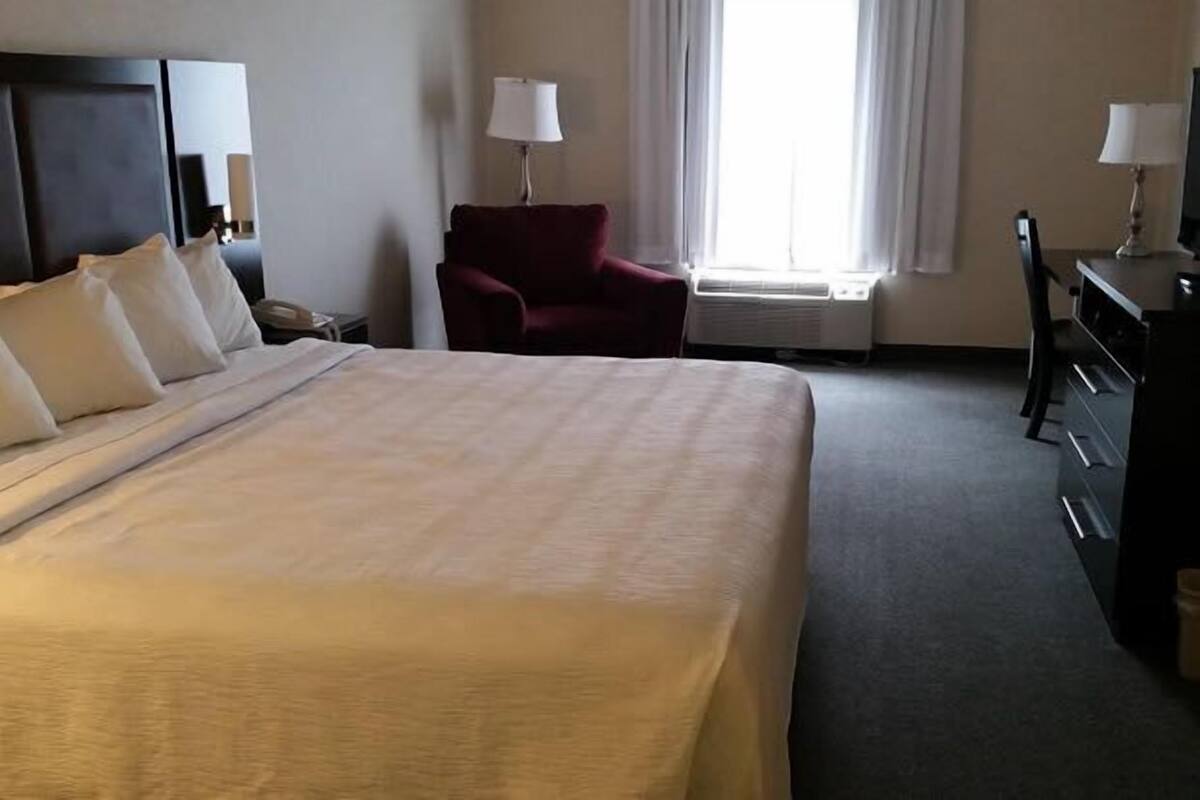 Una habitación de hotel con una cama grande, un sillón rojo, un escritorio con lámpara y una ventana con cortinas.