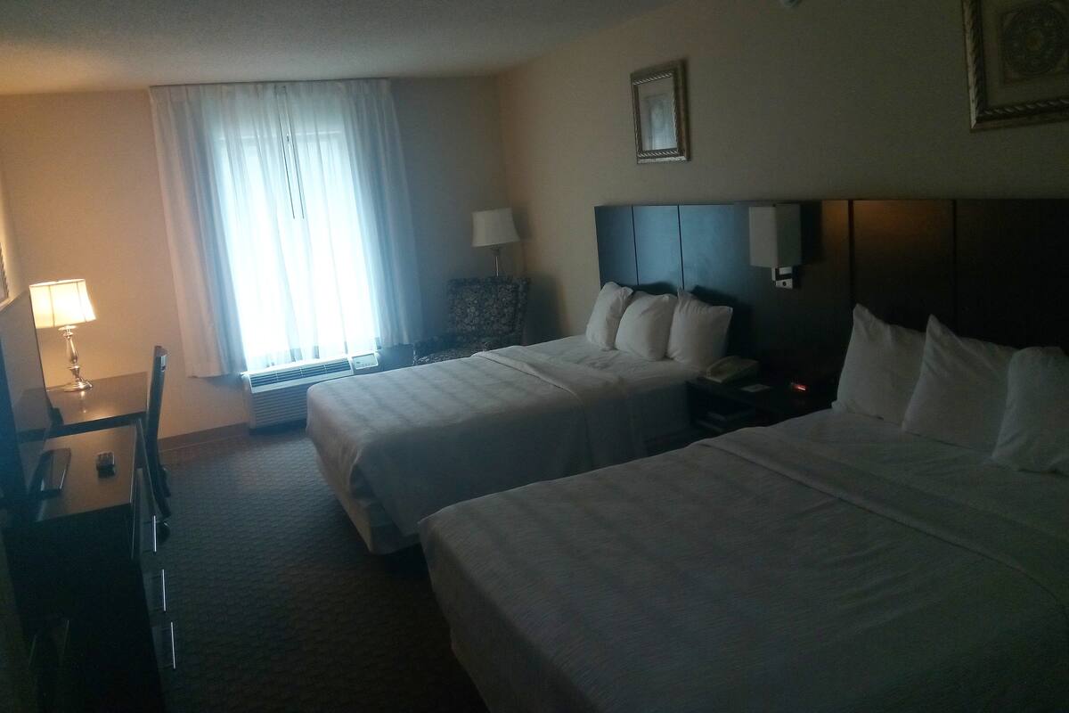 Habitación de hotel con dos camas, un escritorio con lámpara, una silla y un ventanal con cortinas.