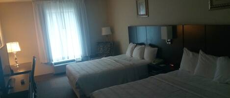 A hotel room with two beds, a desk with a lamp, a chair, and a window with curtains.