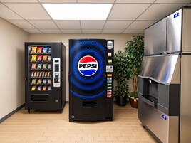 Una máquina expendedora con el logotipo de Pepsi, un refrigerador y una planta en maceta en un espacio interior.