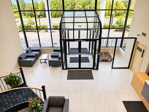 A modern lobby with a glass elevator, seating area, and a staircase with a railing. - Gold Vault Inn (Radcliff)